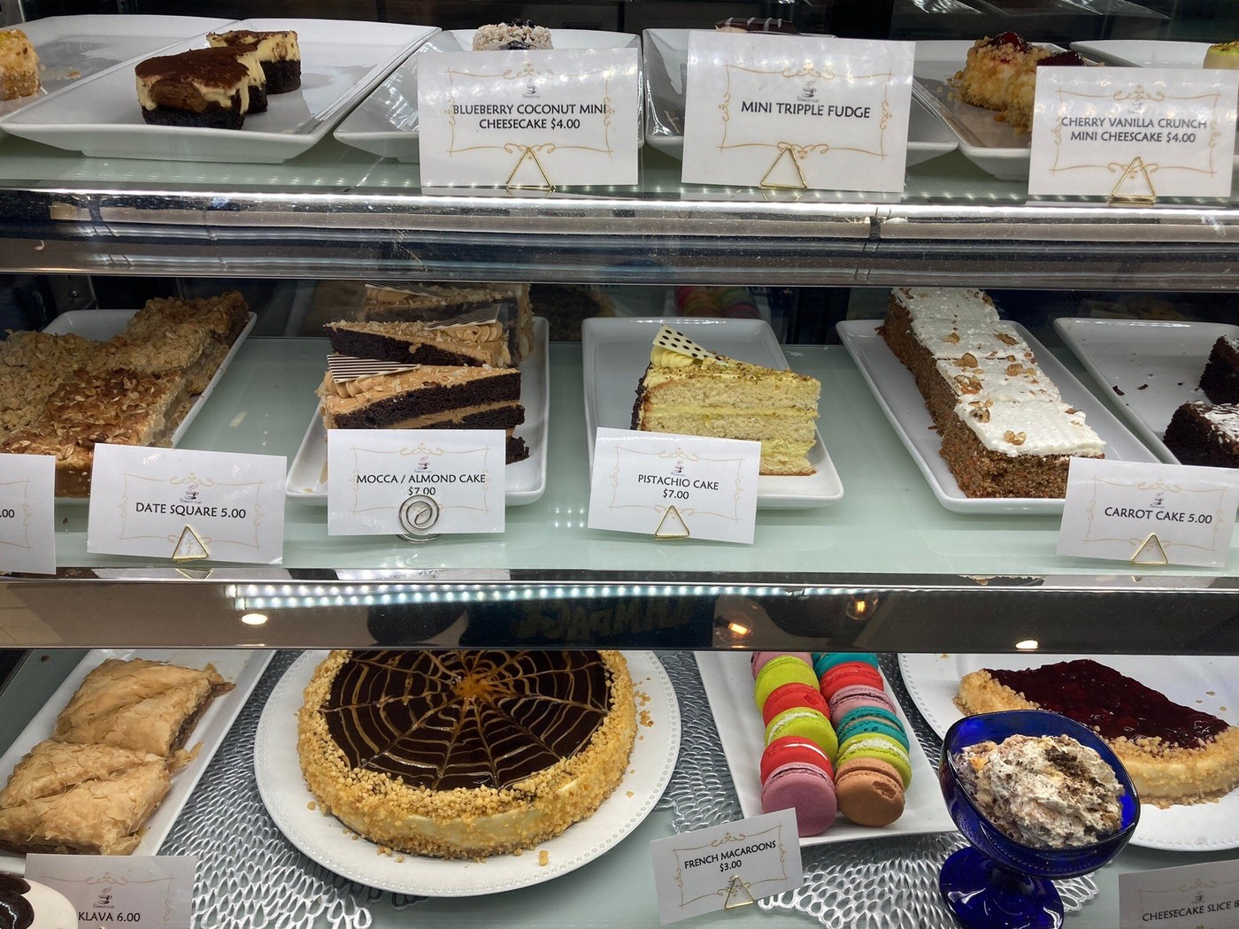 Glass dessert display with individual cakes, layered slices, pastries and colourful macarons on white trays.