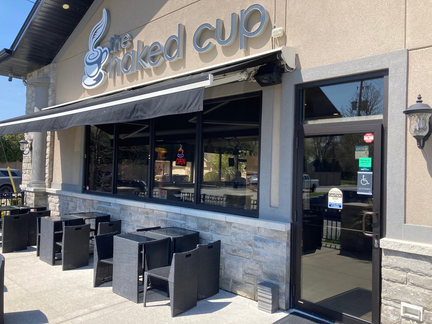 The Naked Cup storefront with raised lettering, a black awning and outdoor tables and chairs.