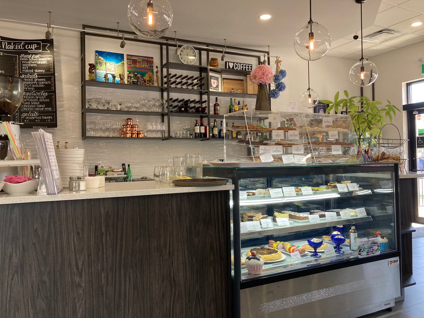 Glass dessert display beneath globe pendant lights, with shelves, artwork, flowers and stacked plates around the café counter.